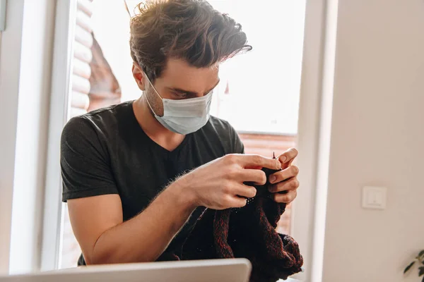 Giovane uomo in maschera medica maglieria vicino al computer portatile a casa — Foto stock