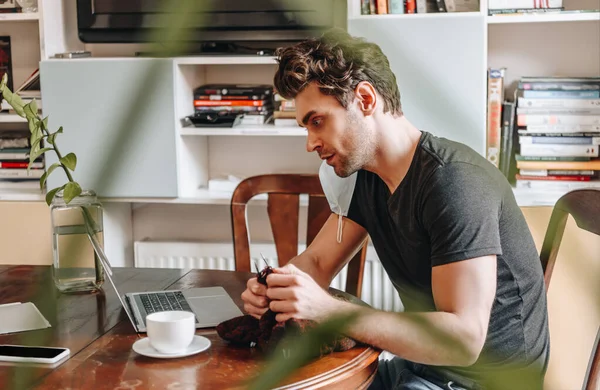 Vista laterale del giovane concentrato che guarda il computer portatile mentre lavora a maglia a casa — Foto stock