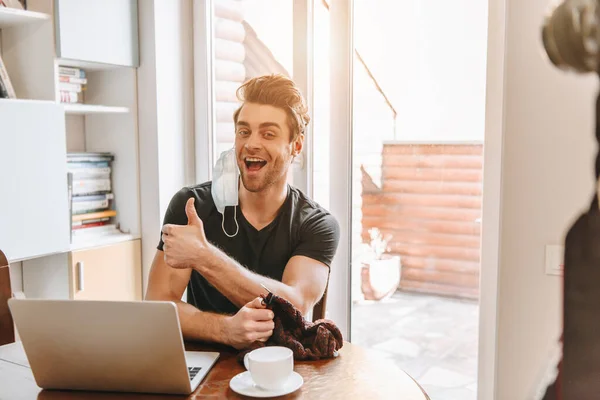 Excited vlogger with medical mask on ear showing thumb up while holding knitting threads — Stock Photo