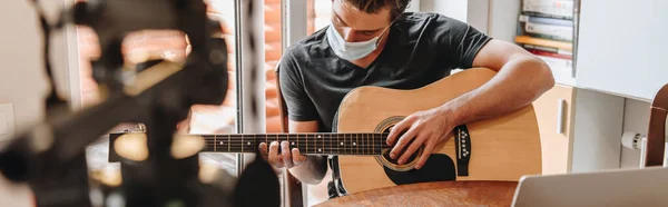 Selective focus of young vlogger in protective mask playing guitar near digital camera on tripod, horizontal image — Stock Photo