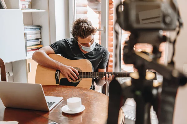 Foyer sélectif du jeune vlogger dans le masque de protection jouer de la guitare près de l'ordinateur portable et appareil photo numérique sur trépied — Photo de stock