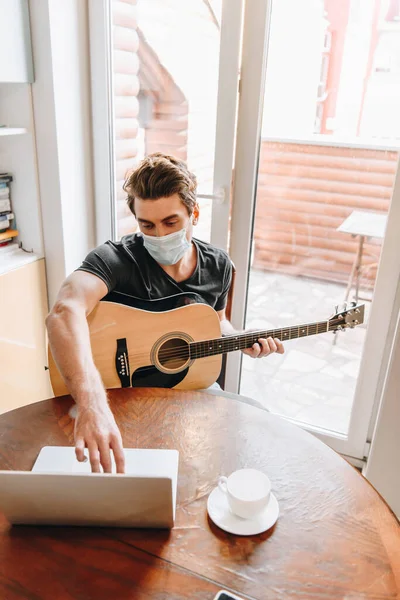 Высокий угол зрения человека в медицинской маске с гитарой и с помощью ноутбука дома — стоковое фото