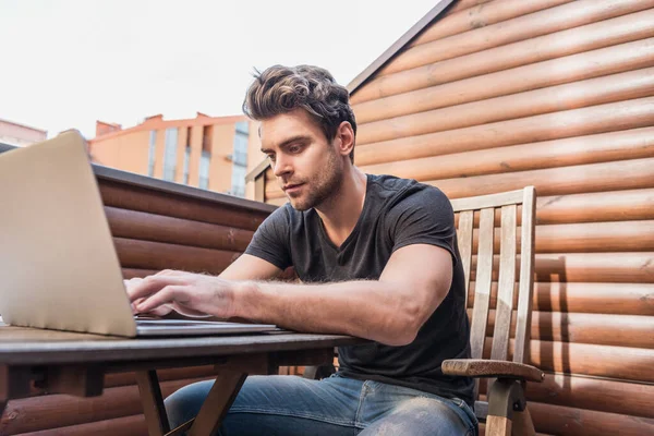 Gutaussehender, konzentrierter Mann tippt auf Laptop, während er auf Balkon sitzt — Stockfoto