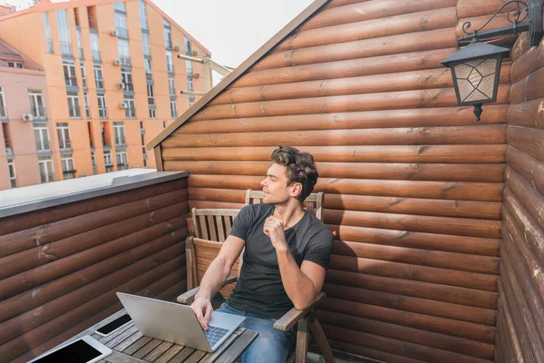 Junger, gutaussehender Mann schaut weg, während er auf dem Balkon in der Nähe des Laptops sitzt — Stockfoto