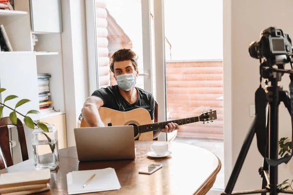 Giovane video blogger in maschera medica che tiene la chitarra mentre è seduto vicino al computer portatile e alla fotocamera digitale su treppiede — Foto stock