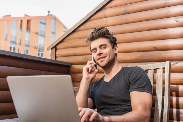 Hombre guapo usando el ordenador portátil y hablando en el teléfono inteligente mientras está sentado en el balcón y sonriendo a la cámara — Stock Photo