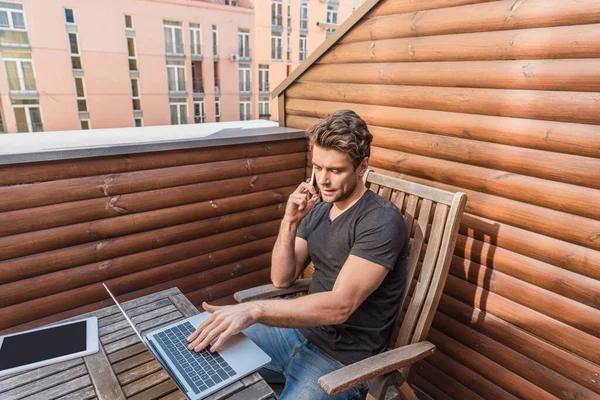 Guapo, hombre serio usando el ordenador portátil y hablando en el teléfono inteligente mientras está sentado en el balcón — Stock Photo
