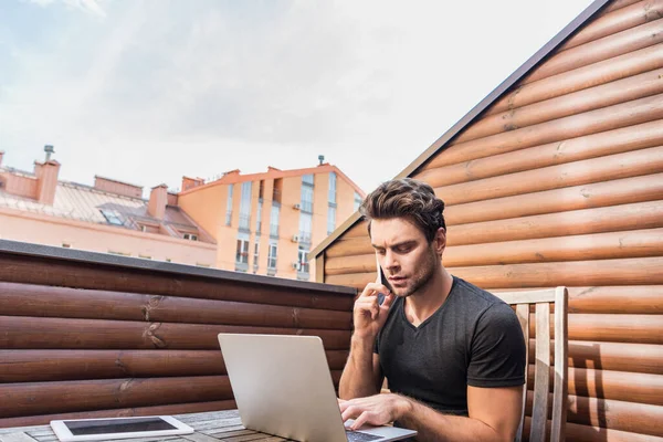 Joven, hombre serio usando el ordenador portátil y hablando en el teléfono inteligente mientras está sentado en el balcón - foto de stock