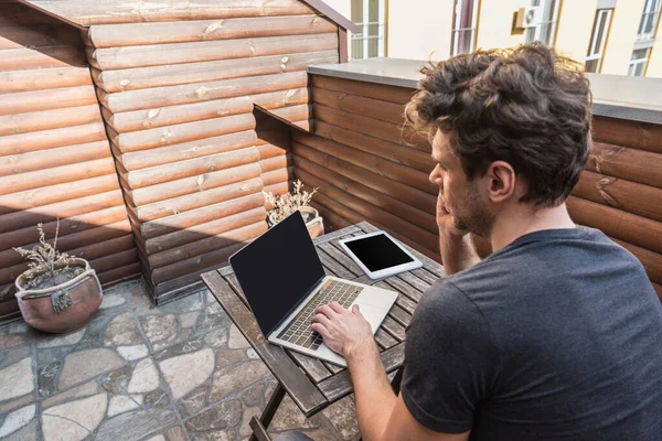 Hombre joven que usa el ordenador portátil con pantalla en blanco cerca de la tableta digital mientras está sentado en el balcón - foto de stock