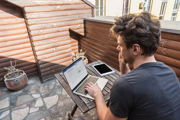 KYIV, UKRAINE - 13 AVRIL 2019 : jeune homme utilisant un ordinateur portable avec le site Facebook assis sur le balcon — Photo de stock