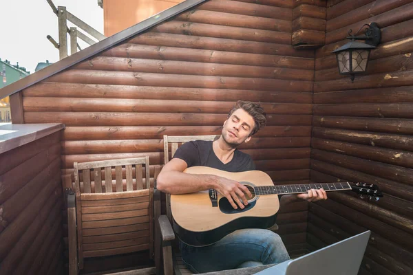 Homme rêveur jouer de la guitare tout en étant assis sur le balcon près d'un ordinateur portable — Photo de stock