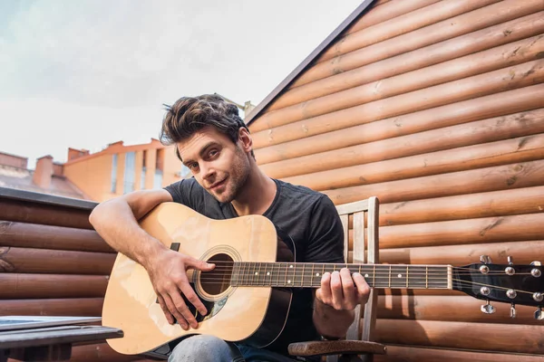 Smiling, handsome man looking at camera while sitting on balcony and playing guitar — Stock Photo