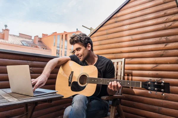 Красивий молодий чоловік тримає гітару і використовує ноутбук, сидячи на балконі — стокове фото