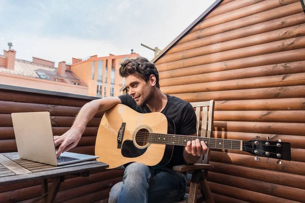 Bello giovane uomo che tiene la chitarra e utilizzando il computer portatile mentre seduto sul balcone — Foto stock