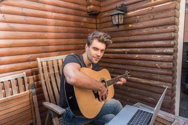 Schöner Mann schaut in die Kamera, während er auf dem Balkon neben Laptop sitzt und Gitarre spielt — Stockfoto