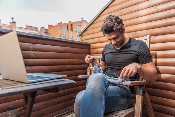 Hombre guapo y sonriente tejiendo mientras está sentado en el balcón cerca de la computadora portátil — Stock Photo