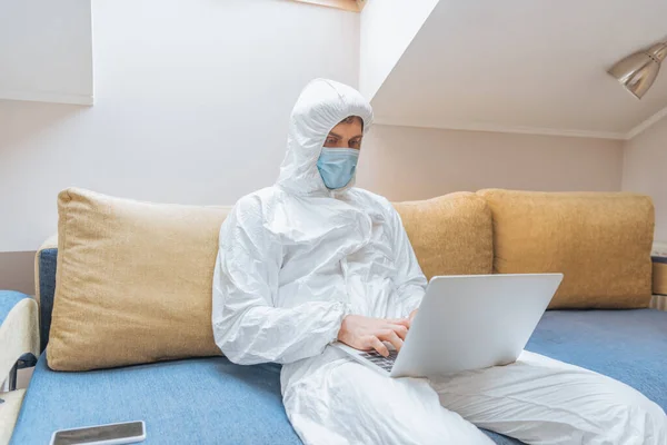 Young man in hazmat suit and protective mask sitting on sofa near smartphone with blank screen and using laptop — Stock Photo