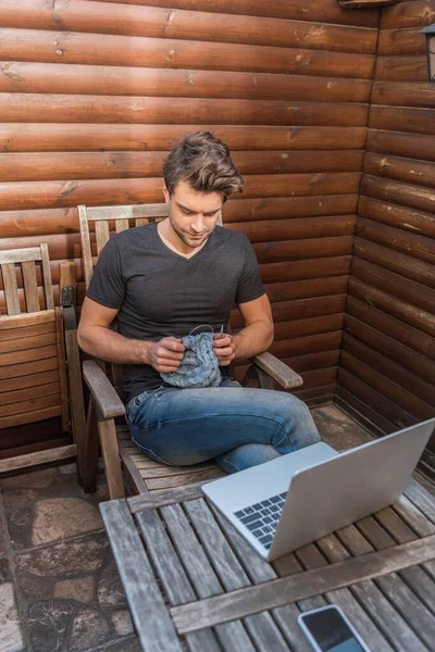 Blick aus der Vogelperspektive auf einen hübschen jungen Mann, der strickt, während er neben dem Laptop auf dem Balkon sitzt — Stockfoto