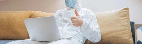 Cropped view of man in hazmat suit and protective mask showing thumb up while sitting on sofa and using laptop, panoramic shot — Stock Photo