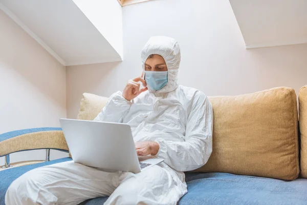 Hombre joven en traje de materiales peligrosos y máscara protectora usando el ordenador portátil y hablando en el teléfono inteligente mientras está sentado en el sofá - foto de stock