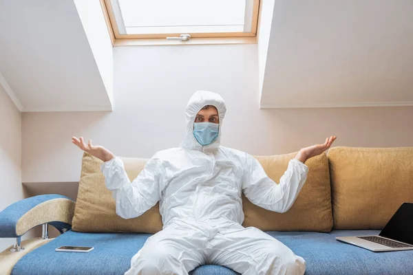 Hombre confuso en traje de materiales peligrosos y máscara protectora sentado en el sofá y mostrando gesto de encogimiento cerca de la computadora portátil y el teléfono inteligente - foto de stock