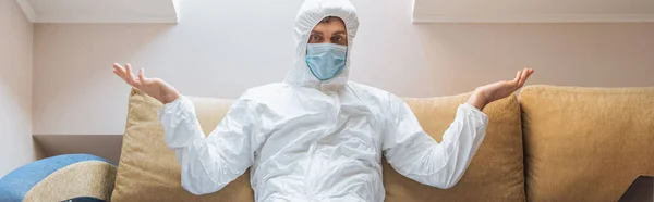 Immagine orizzontale di uomo confuso in tuta hazmat e maschera protettiva seduto sul divano e mostrando gesto scrollata di spalle — Foto stock