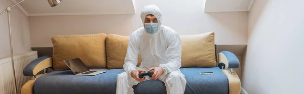 KYIV, UKRAINE - APRIL 13, 2019: panoramic shot of young man in hazmat suit and protective mask sitting on sofa near laptop and looking at camera — Stock Photo