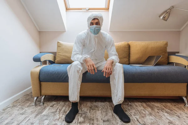 Young man in hazmat suit and protective mask sitting on sofa near laptop and looking at camera — Stock Photo