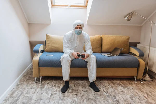 KYIV, UKRAINE - APRIL 13, 2019: young man in hazmat suit and protective mask playing video game while sitting on sofa near laptop and smartphone — Stock Photo