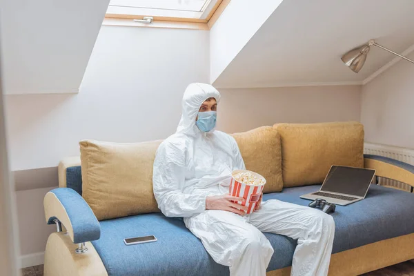 KYIV, UKRAINE - 13 AVRIL 2019 : Un homme en combinaison et un masque de protection tenant un seau de maïs soufflé assis sur un canapé près d'un ordinateur portable, d'un joystick et d'un smartphone — Photo de stock