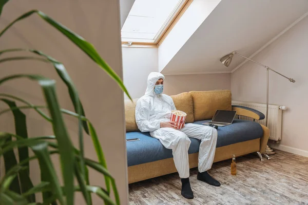 Selective focus of man in hazmat suit and protective mask sitting on sofa near laptop while holding popcorn bucket — Stock Photo