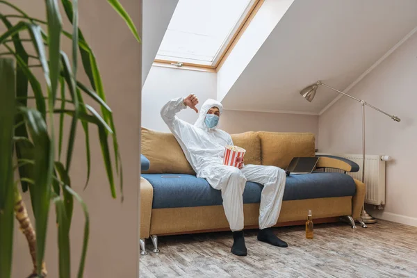 Selective focus of man in hazmat suit and protective mask sitting on sofa near laptop, holding popcorn bucket and showing thumb down — Stock Photo