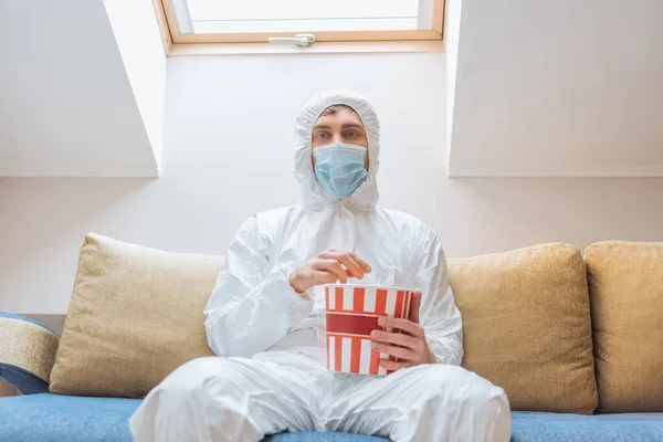 Hombre joven en traje de materiales peligrosos y máscara protectora sentado en el sofá con palomitas de maíz cubo y mirando a la cámara - foto de stock