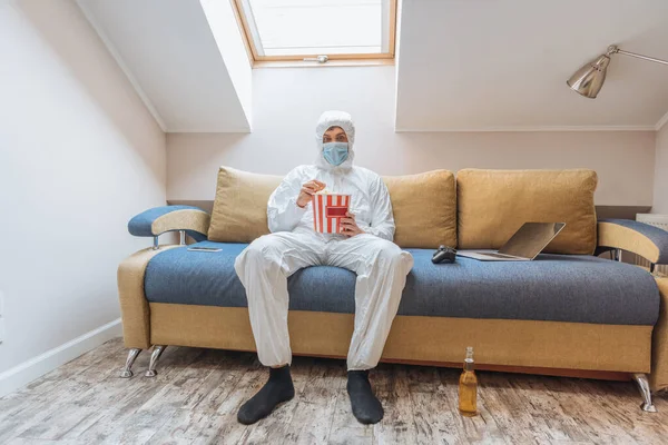 KYIV, UKRAINE - APRIL 13, 2019: young man in hazmat suit and protective mask sitting on sofa and eating popcorn near joystick and laptop — Stock Photo