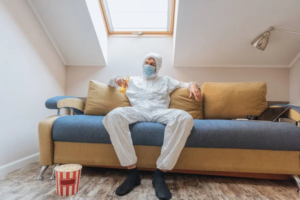 Jeune homme en combinaison Hazmat et masque de protection tenant bouteille de bière tout en étant assis sur le canapé près seau de maïs soufflé sur le sol — Photo de stock