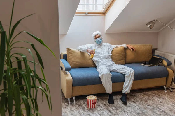 Jeune homme en combinaison Hazmat et masque de protection tenant bouteille de bière tout en étant assis sur le canapé près seau de maïs soufflé sur le sol — Photo de stock