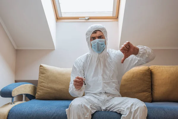 Displeased man in hazmat suit and protective mask showing thumb down while holding thermometer — Stock Photo