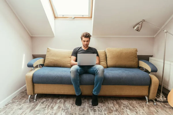 Schöner, aufmerksamer junger Mann sitzt mit Laptop auf Sofa im Dachgeschoss — Stockfoto