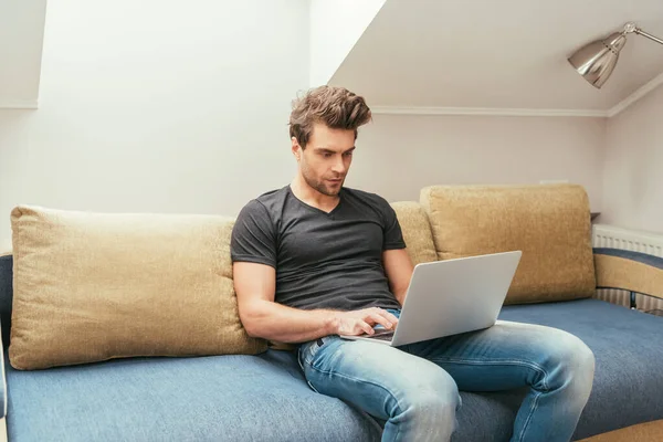 Atento, joven guapo usando el ordenador portátil mientras está sentado en el sofá en casa - foto de stock