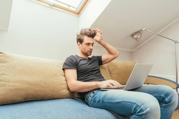 Nachdenklicher junger Mann berührt Kopf während er zu Hause am Laptop arbeitet — Stockfoto