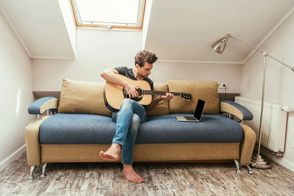 Joven tocando la guitarra mientras está sentado en el sofá y mirando el portátil con pantalla en blanco - foto de stock