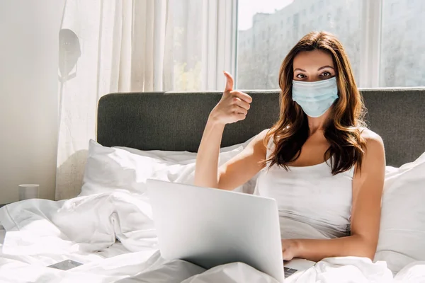 Junge Freiberuflerin mit medizinischer Maske zeigt Daumen nach oben, während sie während der Selbstisolierung im Bett am Laptop arbeitet — Stockfoto