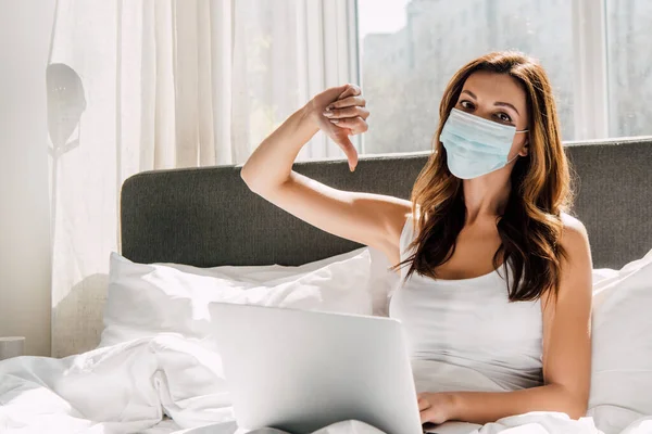 Фрілансерка в медичній масці показує великий палець вниз під час роботи на ноутбуці під час самоізоляції в ліжку — стокове фото