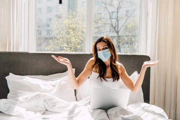 Female freelancer in medical mask with shrug gesture working on laptop during self isolation in bed — Stock Photo