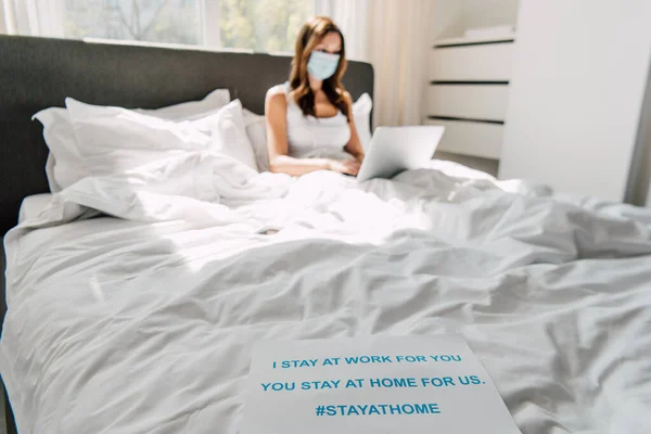 Young freelancer in medical mask working on laptop in bed with Stay at home banner during self isolation, selective focus — Stock Photo