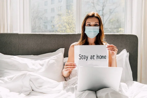 Freiberuflerin in medizinischer Maske mit Bleibe zu Hause-Schild und Arbeit am Laptop während der Selbstisolierung im Bett — Stockfoto