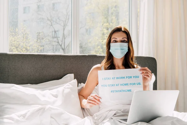 Young freelancer in medical mask holding Stay at home banner and working on laptop during self isolation in bed — Stock Photo