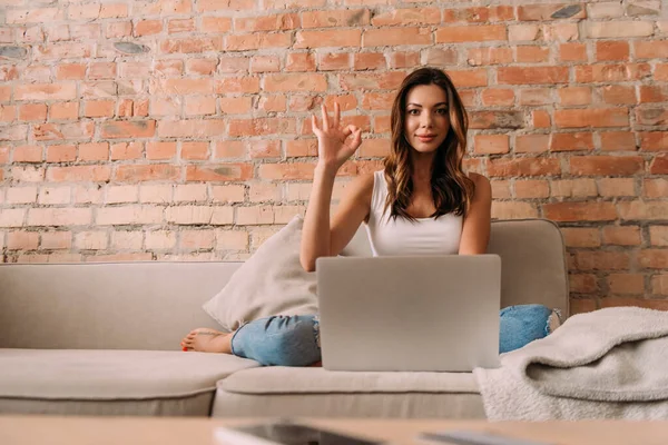 Attraktive Freiberuflerin, die während der Arbeit am Laptop auf dem Sofa während der Selbstisolierung OK-Zeichen zeigt — Stockfoto