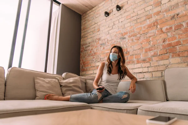 Frau in medizinischer Maske hält Fernbedienung in der Hand und schaut während der Selbstisolierung auf dem Sofa fern — Stockfoto