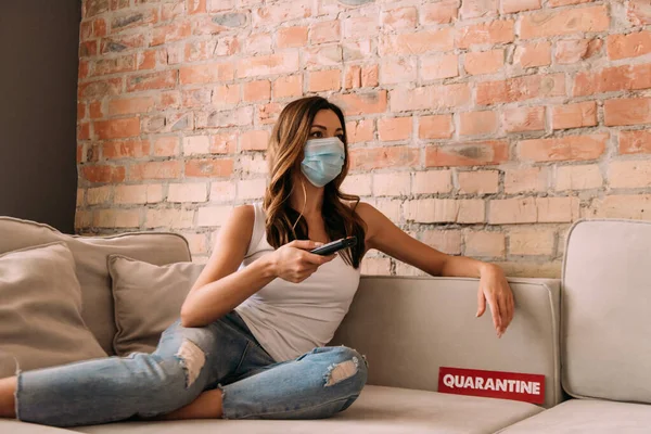 Jolie fille dans le masque médical tenant télécommande et regarder la télévision sur le canapé avec signe de quarantaine — Photo de stock